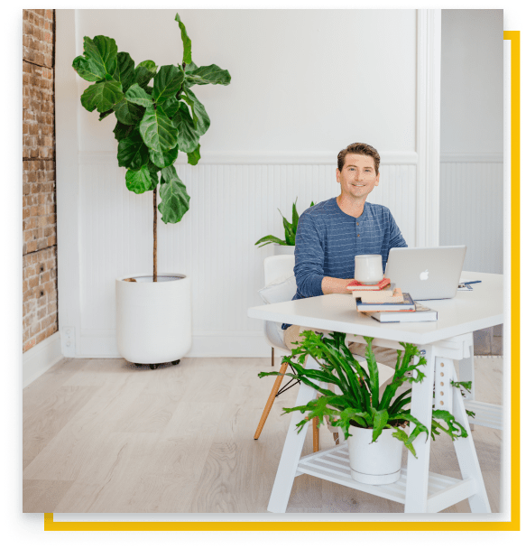 Greg Gall sits at a table that is to the right of the image. A large plant is behind him to the left.