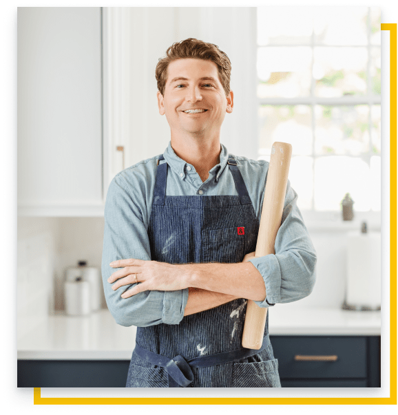 Greg holds a rolling pin and wears an apron standing in a kitchen smiling.